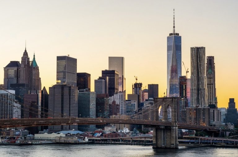 Sunset view of the New York City skyline showcasing the Brooklyn Bridge in the foreground and the One World Trade Center in the background, with various other skyscrapers and the Hudson River.