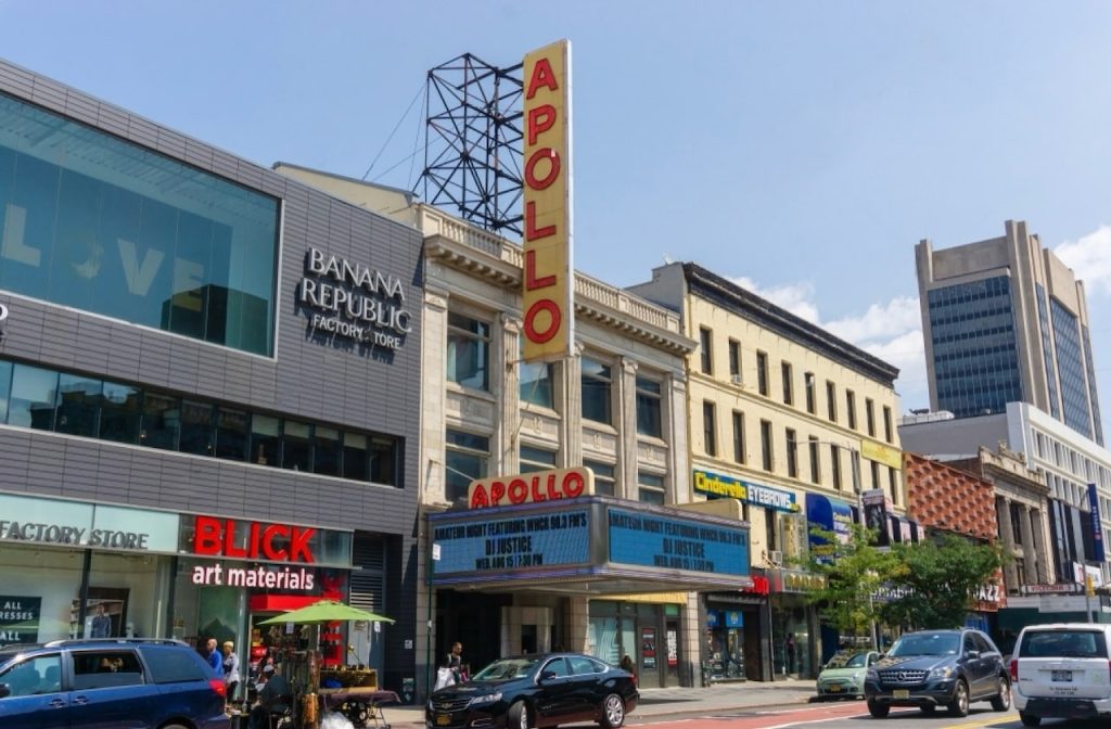 24 hours in NYC: The iconic Apollo Theater marquee stands out on a busy Harlem street with a mix of contemporary and historic buildings, alongside retail stores like Banana Republic and Blick Art Materials, under a clear sky.