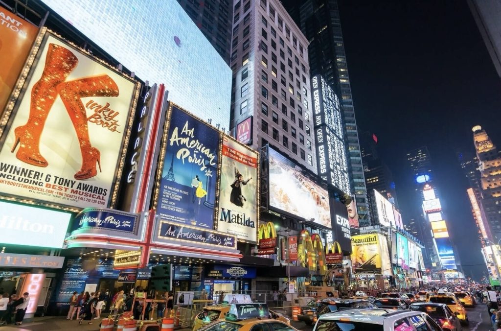 24 hours in NYC: Night time in Times Square with bright billboards for Broadway shows like 'Kinky Boots', 'An American in Paris', and 'Matilda' illuminating the bustling street filled with taxis and crowds.