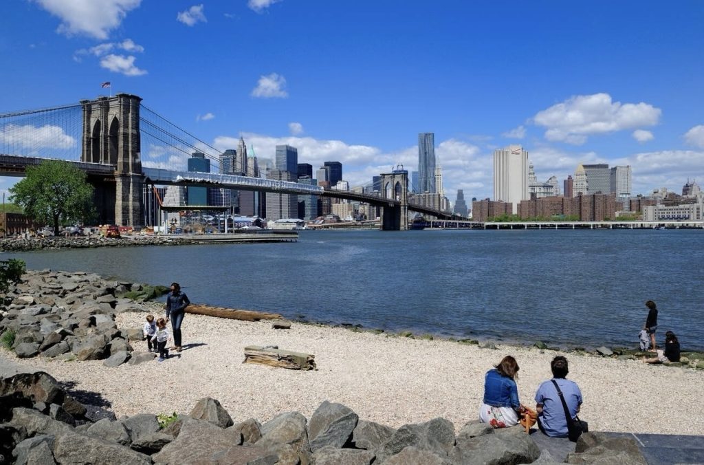 A clear day at Brooklyn Park with visitors enjoying the view of the Brooklyn Bridge and the Manhattan skyline across the East River, featuring a mix of clouds and blue sky overhead.