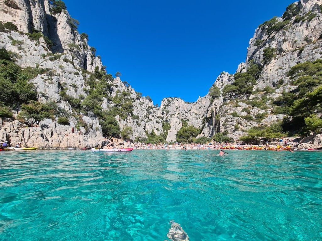 Breathtaking view of Calanque d'En-Vau near Marseille, with its crystal-clear turquoise waters gently lapping against a pebbled beach, framed by towering rugged limestone cliffs under a clear blue sky. The scene is vibrant with the summer buzz of beachgoers enjoying the sun and the sea.