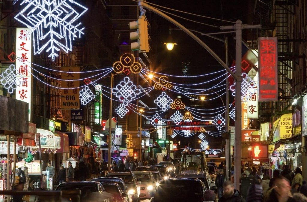 24 hours in NYC: Evening view of a lively street in Chinatown, New York City, adorned with illuminated snowflake decorations and a vibrant display of neon signs, bustling with pedestrians and traffic.