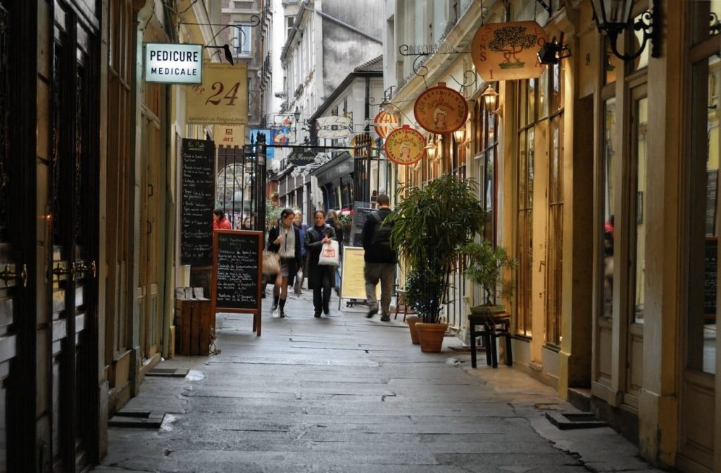 Prettiest Streets in Paris: Shoppers and diners navigate the historic Cour du Commerce Saint-André in Paris, characterized by its old stone pavement and classic shop signs, including one for 'Pedicure Medicale.' The narrow passageway is framed by golden-hued storefronts offering a variety of wares and services, with chalkboard menus and green potted plants inviting passersby to indulge in the area's rich culinary and cultural offerings.