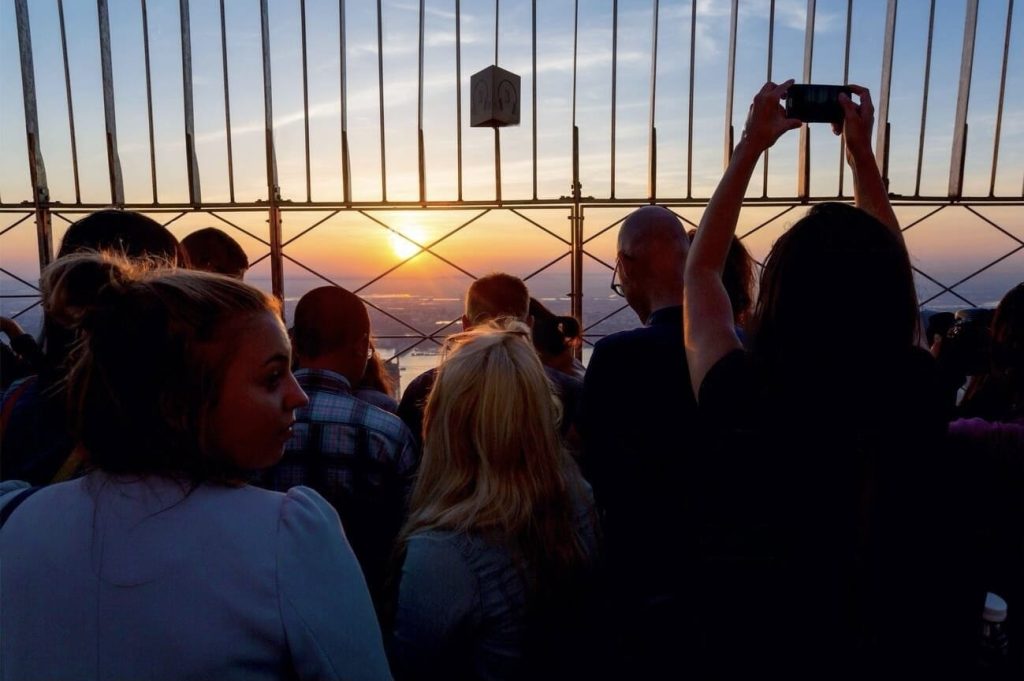 24 hours in NYC: Silhouettes of people gathered at an observation deck, captured against the backdrop of a stunning sunset with vibrant hues in the sky, with one individual taking a photo to capture the moment.