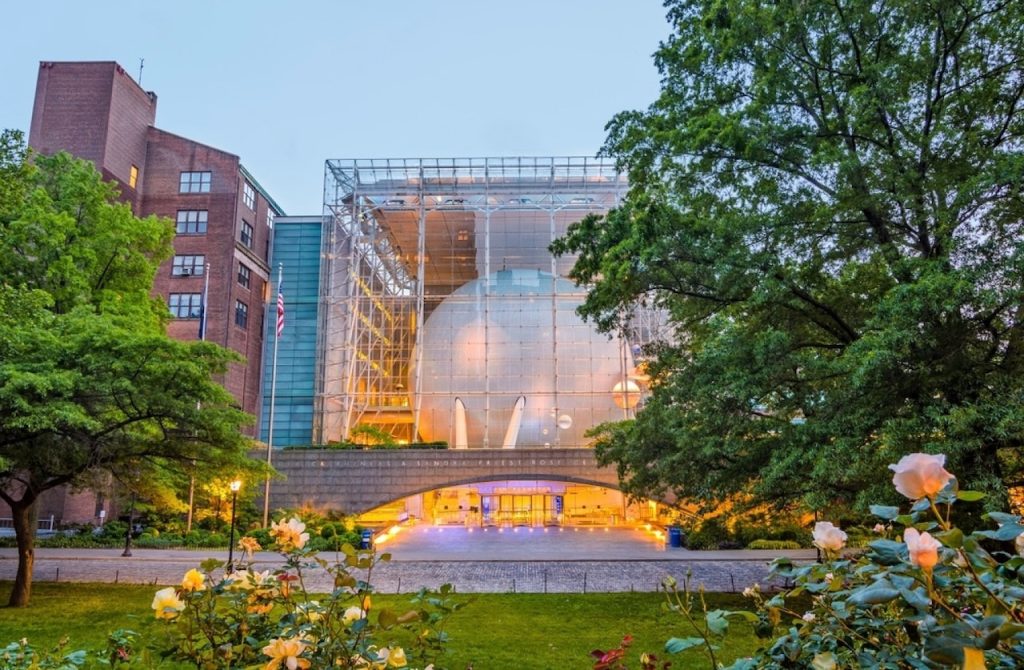 24 hours in NYC: Twilight view of the American Museum of Natural History's Rose Center for Earth and Space, with the transparent glass cube revealing the spherical Hayden Planetarium, surrounded by lush greenery and blooming roses in the foreground.