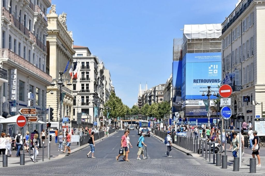 A bustling urban street in Marseille with pedestrians crossing and walking along the sidewalk. The architecture lining the street is quintessentially European, featuring facades with intricate details and French flags. In the background, construction scaffolding is draped with a large blue banner, adding a contemporary touch to the historic city atmosphere.