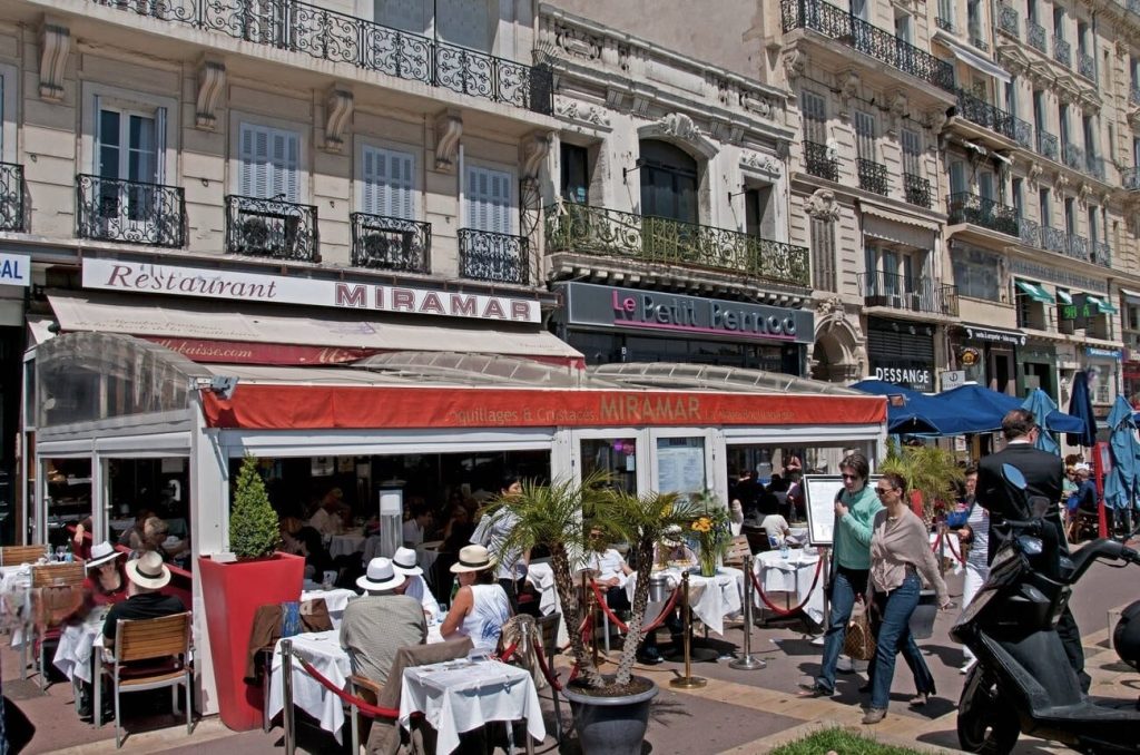 Busy outdoor dining scene at Restaurant Miramar on a lively street in Marseille, with patrons enjoying meals under red awnings. Elegant white tablecloths and casual, fashionable attire of diners capture the relaxed yet sophisticated atmosphere of the city. The street buzzes with activity, including pedestrians and a parked motor scooter, against the backdrop of classic French architecture with ornate balconies.