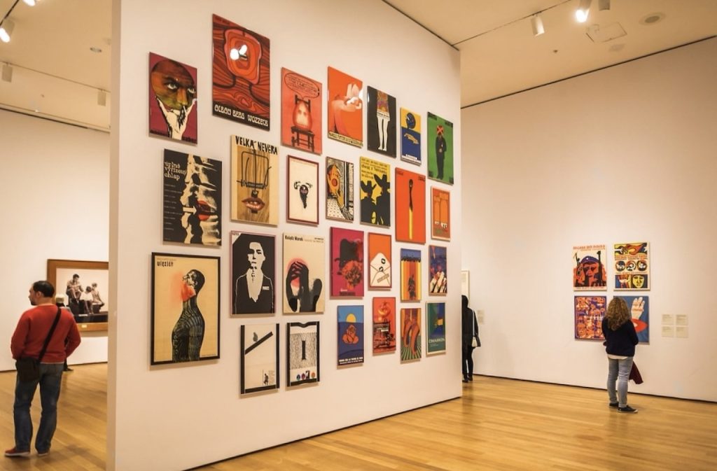 Visitors admiring a colorful collection of modern graphic posters on a white wall at the Museum of Modern Art (MoMA) in New York City, reflecting the diverse styles and subjects of contemporary art.