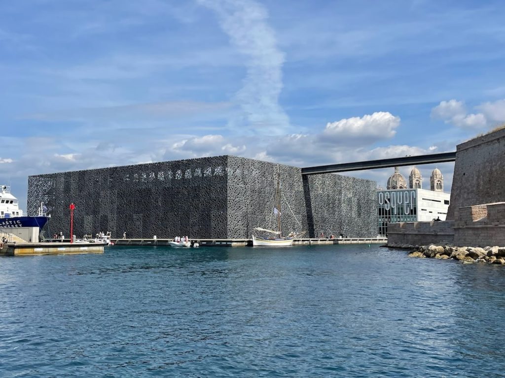 The Museum of European and Mediterranean Civilisations (MuCEM) in Marseille, France, stands out with its modern, intricate lattice cube structure, juxtaposed against the historical backdrop of the old port. The museum is connected by a sleek footbridge to the neighboring Fort Saint-Jean, displaying a harmonious blend of contemporary architecture and ancient heritage against the serene blue of the Mediterranean Sea.