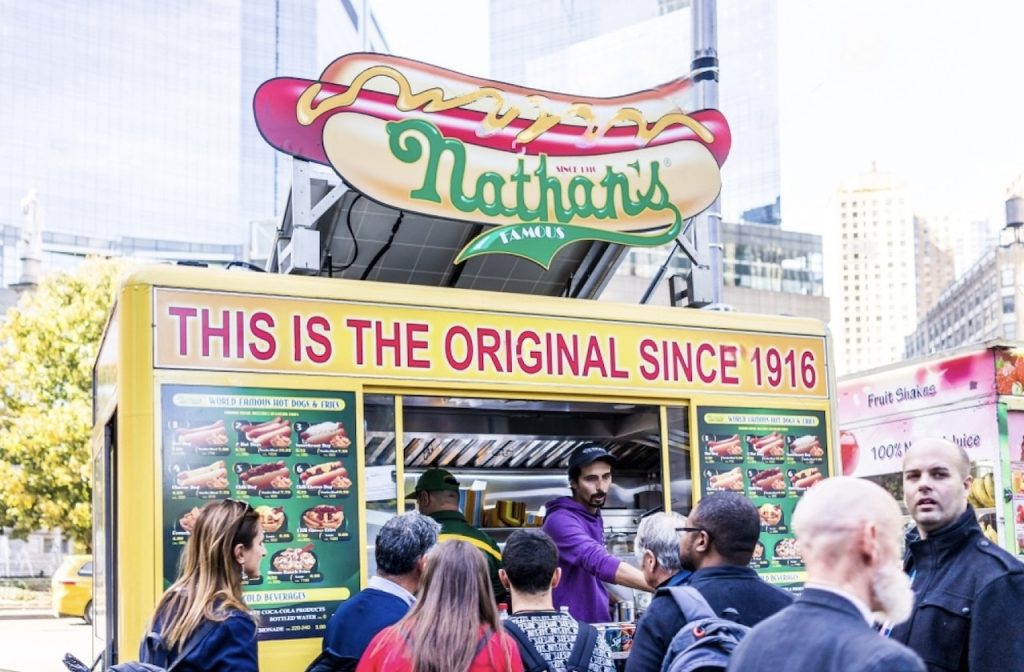 Queue of customers at Nathan's Famous hot dog stand, with a prominent sign stating 'THIS IS THE ORIGINAL SINCE 1916', highlighting the brand's long history of serving hot dogs and fries in New York City.