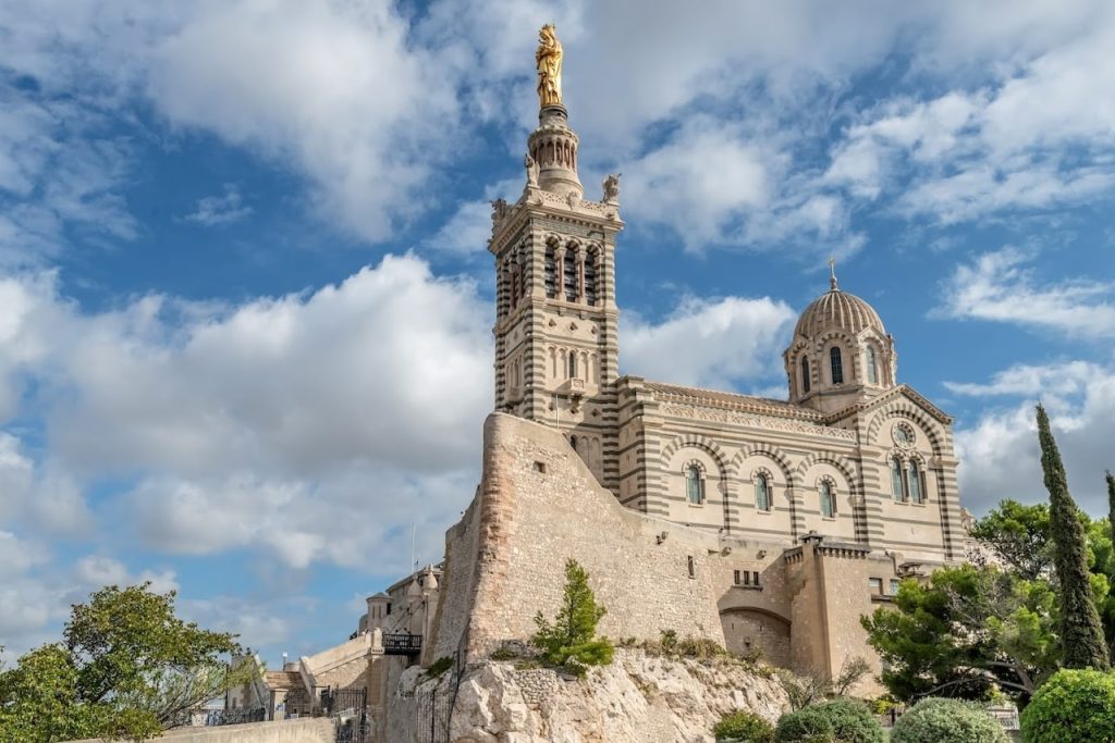 The majestic Notre-Dame de la Garde basilica stands atop a limestone outcrop overlooking Marseille, its ornate neo-Byzantine architecture crowned with a gilded statue of the Virgin Mary. The basilica, with its striped stone facade, is a beacon of faith and a historical landmark, set against a dynamic sky, symbolizing its watchful presence over the city.