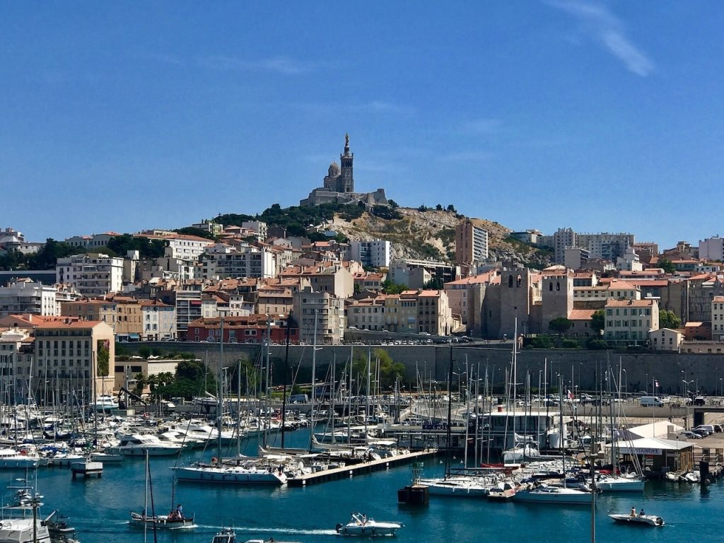 Scenic view of the old port of Marseille, Vieux-Port, bustling with an array of sailboats and yachts moored in the marina. The iconic Notre-Dame de la Garde basilica sits atop the hill, overlooking the dense urban landscape of terracotta-roofed buildings and the tranquil blue waters of the Mediterranean Sea, under a clear sky.