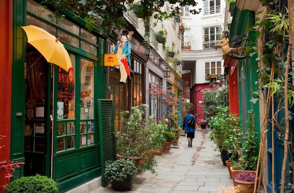 Prettiest Streets in Paris: A narrow alleyway, Passage de l'Ancre in Paris, offers a charming and colorful stroll with its vibrant shopfronts and whimsical hanging signs, including one with a figure holding an umbrella. Lined with potted plants and climbing vines, the passage exudes a cozy, hidden-garden feel as a lone individual walks away, adding a sense of quiet daily life to this enchanting urban retreat.