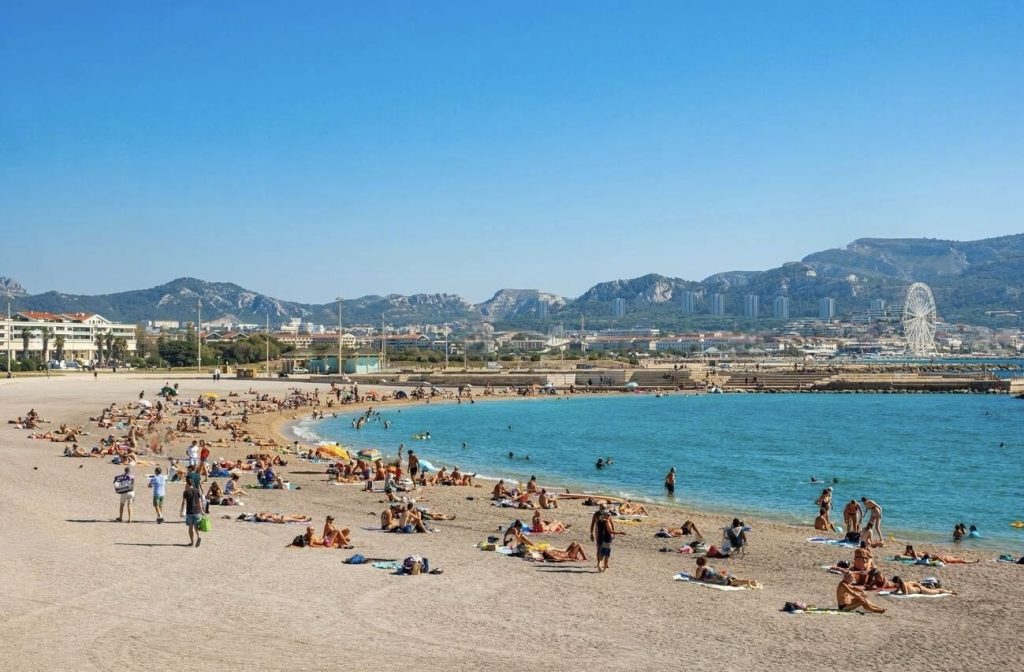 Sun-seekers bask on the sandy expanse of Prado Beaches in Marseille, set against a panoramic view of distant mountains and urban skyline featuring a Ferris wheel. The Mediterranean Sea's azure waters gently kiss the shore, while the city's lively atmosphere is hinted at by the bustling beachfront and the relaxed, leisurely vibe of the beachgoers.