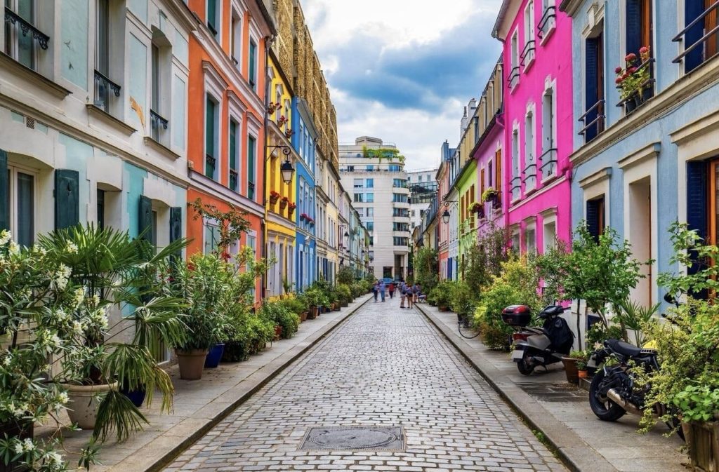 Prettiest Streets in Paris: A charming cobblestone street lined with colorful houses in varying shades of orange, blue, pink, and green, with potted plants and trees adding greenery to the urban scene. People are visible walking in the distance, enhancing the lively yet quaint atmosphere of the neighborhood.