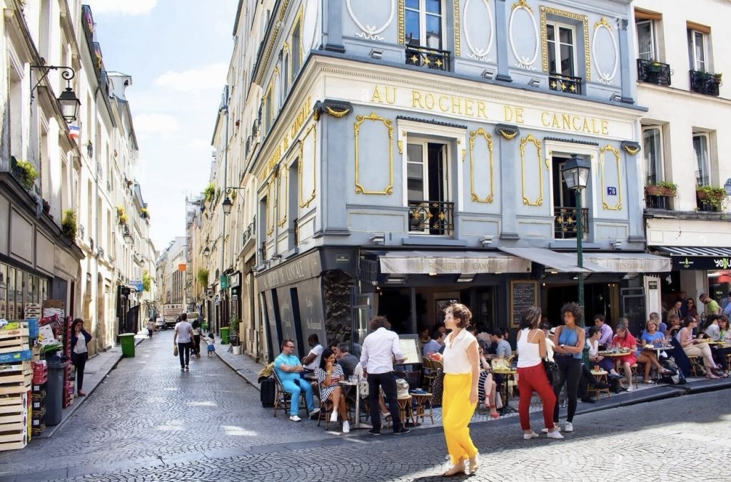 A vibrant city street scene featuring the corner of 'Au Rocher de Cancale', a restaurant with an elegant blue and gold façade. People are seated outside at bistro tables enjoying the ambiance, while others walk along the cobblestone street lined with traditional buildings and shops. The casual bustle of urban life is captured under the bright sunlight, with foliage hanging from the windows above.