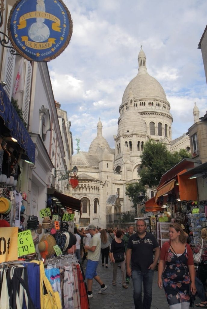 Prettiest Streets in Paris: A bustling street market scene with the iconic Sacré-Cœur Basilica in the background, under a partly cloudy sky. The foreground features a variety of merchandise on display, including hats and clothing, with signs showing prices. Shoppers and tourists navigate through the stalls, with a blue and gold sign reading 'Aux Etoiles de Marie' hanging prominently to the left.