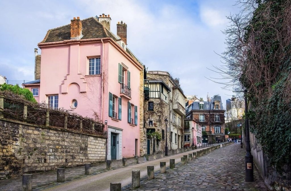 Prettiest Streets in Paris: A quaint cobblestone lane in Montmartre, Paris, with a distinctive pink house at the turn of the street, flanked by traditional buildings with shuttered windows. The historic charm is enriched by the bare vine-covered walls and a lamp post, evoking the artistic and bohemian spirit of the area. The scene is devoid of the typical urban bustle, offering a glimpse into the quieter, more residential side of this famous neighborhood.