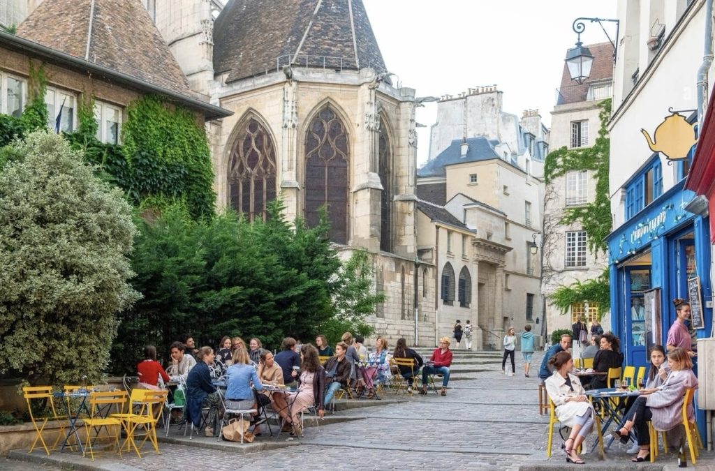 Prettiest Streets in Paris: A cozy outdoor café setting with people seated at yellow and grey tables, engaged in conversation, adjacent to a historical building with gothic-style windows and ivy-covered walls. The scene is nestled within an old European quarter, evidenced by the classic architecture and cobbled streets, with pedestrians milling about in the background, adding to the relaxed and social atmosphere of the location.