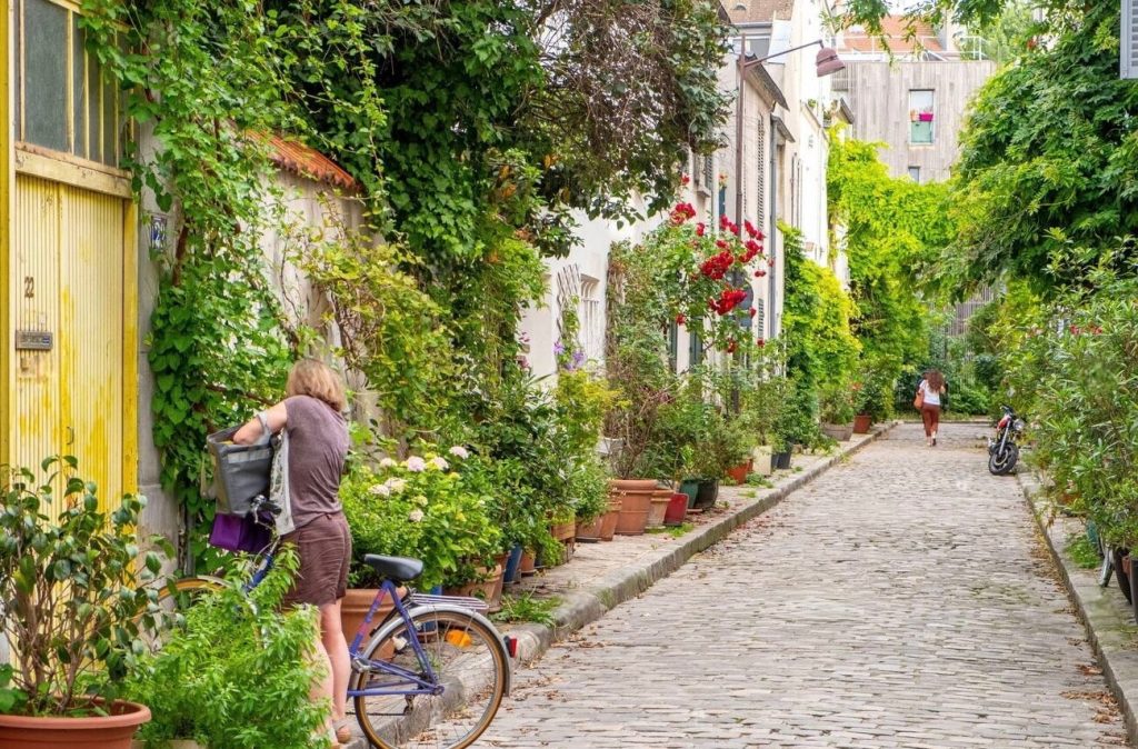 Prettiest Streets in Paris: A person stands with a bicycle on a quaint cobblestone lane, Rue des Thermopyles in Paris, surrounded by lush greenery and flowering plants that spill over garden walls and fences. The rustic charm of the street is accentuated by the vibrant vegetation and colorful facades of the houses. Further down the path, another individual walks away, contributing to the peaceful, everyday life atmosphere of this picturesque urban alley.