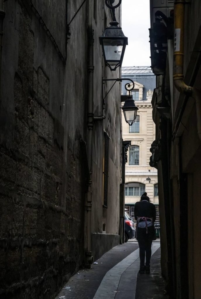 A solitary figure walks through the narrow confines of Rue du Chat-qui-Pêche in Paris, France, known as the narrowest street in the city. The historic mood is set by the weathered stone walls and traditional lanterns, casting a soft glow in the dim light. The image captures a moment of quiet solitude amidst the bustling city, with the contrast between the shadowed alleyway and the brighter, more open space at the end.