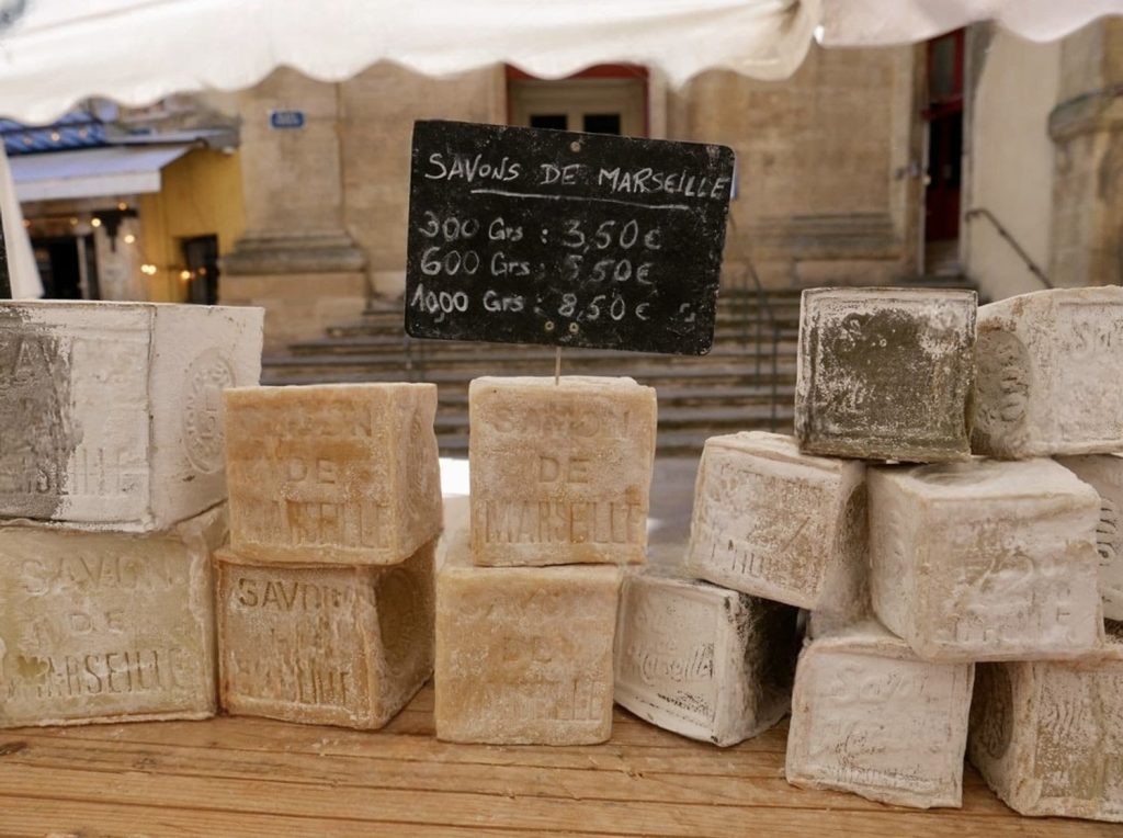 Handcrafted blocks of traditional 'Savon de Marseille' soap displayed on a wooden table, each stamped with the iconic product name. A chalkboard sign lists prices by weight in euros, illustrating the market scene in Marseille where these all-natural, olive oil-based soaps are a local specialty and a popular choice for visitors seeking authentic Provençal goods.