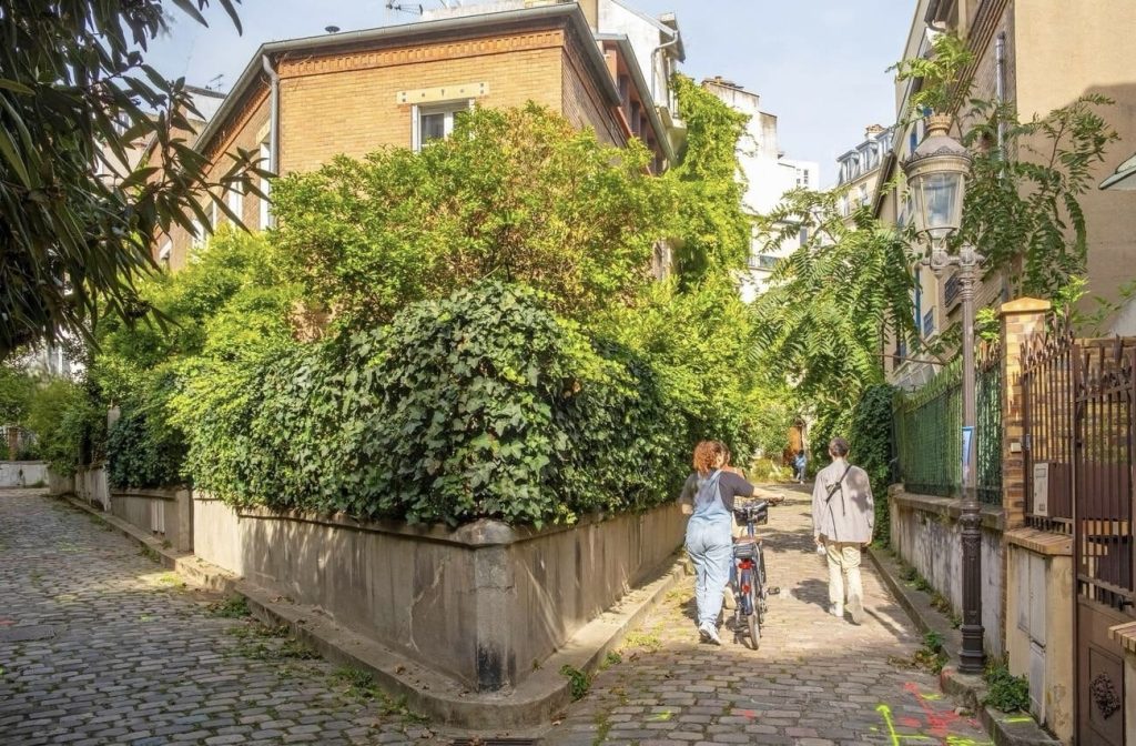 Prettiest Streets in Paris: A peaceful cobbled alleyway shaded by lush overgrown greenery, with a woman walking her bicycle alongside a companion in casual attire. The natural canopy complements the warm brick and stucco façades of the surrounding residential buildings, creating a tranquil urban oasis. Sunlight filters through the leaves, casting a dappled pattern on the path, enhancing the idyllic atmosphere of the scene.