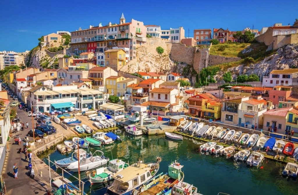 Picturesque view of the small fishing port of Vallon des Auffes in Marseille, with its tightly clustered, colorful houses hugging the hillside and boats moored in the calm blue water. The scene is a vibrant tapestry of Mediterranean architecture, local culture, and maritime heritage, bathed in the warm glow of the sun.