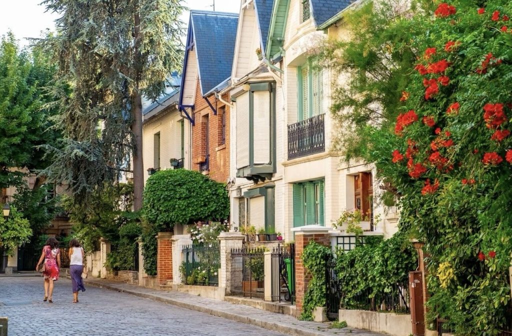 Two people walk down a picturesque street lined with elegant townhouses, featuring a blend of brick and plaster façades and colorful shutters. Lush greenery and bright red flowering bushes add vibrant bursts of color to the tranquil scene, while the cobbled path and manicured gardens evoke a sense of refined calm in this charming urban neighborhood.