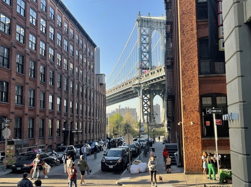 Street-level view in DUMBO, Brooklyn, with pedestrians and parked cars lining the street, flanked by red brick buildings, and the distinctive Manhattan Bridge arching overhead against a clear blue sky.