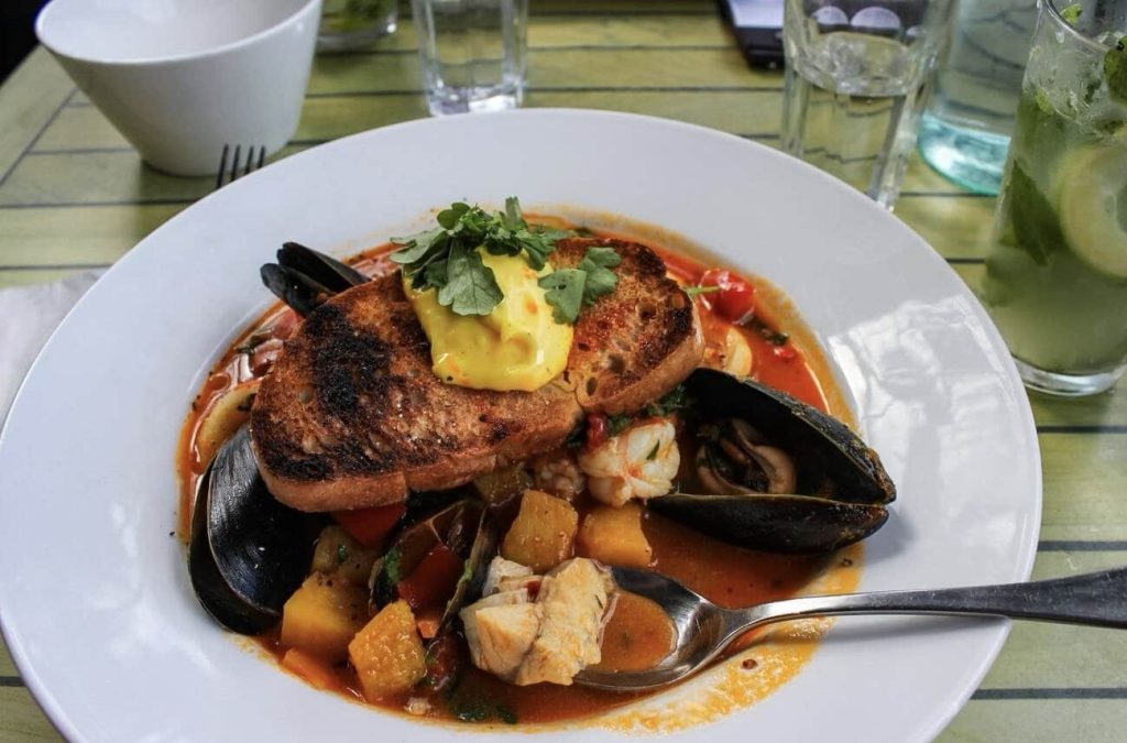 A classic dish of Bouillabaisse, traditional Marseille seafood stew, served in a white bowl. The rich, saffron-infused broth contains a variety of seafood including mussels and fish, accompanied by a toasted slice of bread topped with rouille sauce. A refreshing glass of mojito in the background complements the meal, all set on a rustic green-striped table.