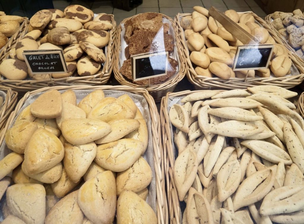 Assorted traditional French biscuits displayed in wicker baskets in a bakery in Marseille, France. The selection includes 'Navettes,' a specialty of the region, which are boat-shaped cookies flavored with orange blossom, alongside 'Galets de Nice,' chocolate and orange cookies. Each type is neatly labeled, showcasing the local culinary delights and artisanal baking craftsmanship.