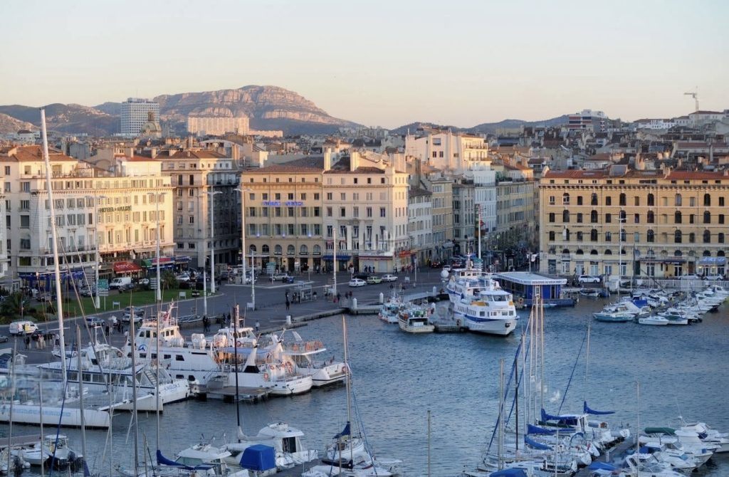 The golden hour casts a warm glow over Vieux-Port in Marseille, where a variety of boats and yachts are moored in the calm harbor waters. The historic port is framed by classic French architecture and busy streets, leading the eye to the majestic limestone massif in the distance, all under a soft dawn sky.