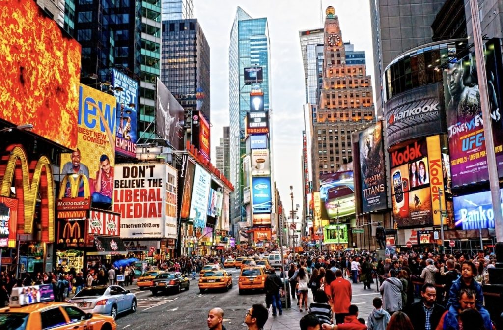 Vibrant and bustling Times Square in New York City, filled with a crowd of pedestrians, yellow taxis, and a multitude of glowing LED billboards advertising Broadway shows, restaurants, and various brands.